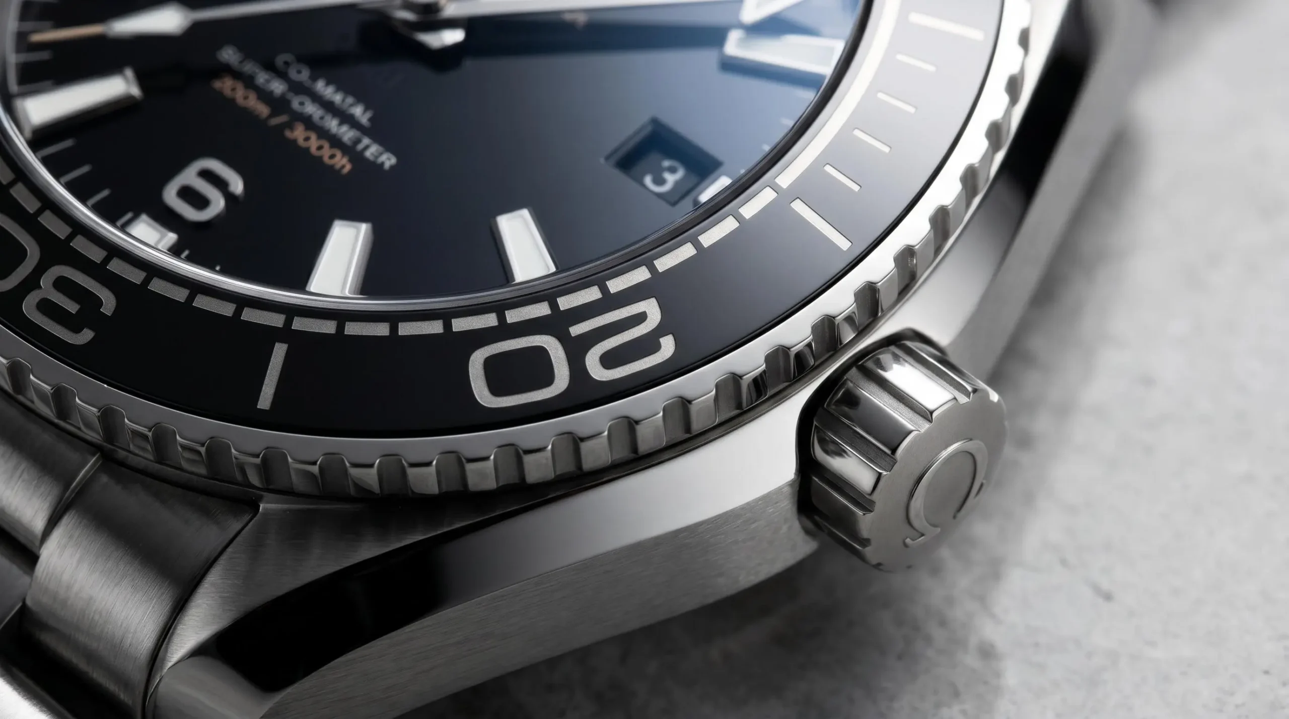 Gros plan macro de la lunette en céramique noire d'une Omega Seamaster Planet Ocean, couronne vissée visible, index luminescents sur cadran noir, reflets de lumière rasante révélant les textures de la céramique mate et de l'acier poli