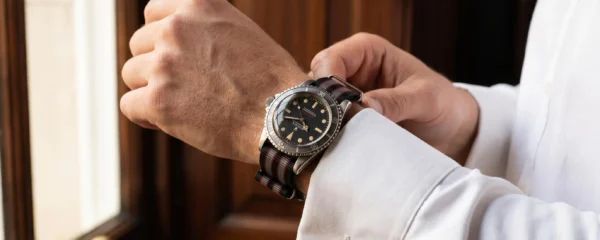 Gros plan d'un poignet masculin portant une Rolex Submariner vintage sur bracelet NATO rayé gris-noir-rouge, manchette de chemise blanche visible, main ajustant un bouton de manchette, intérieur élégant aux boiseries sombres en arrière-plan flou
