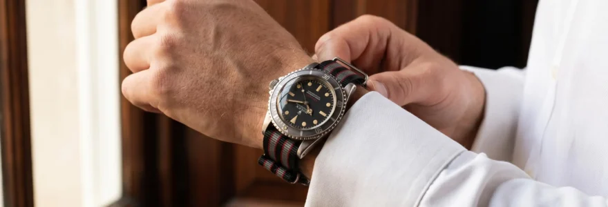 Gros plan d'un poignet masculin portant une Rolex Submariner vintage sur bracelet NATO rayé gris-noir-rouge, manchette de chemise blanche visible, main ajustant un bouton de manchette, intérieur élégant aux boiseries sombres en arrière-plan flou