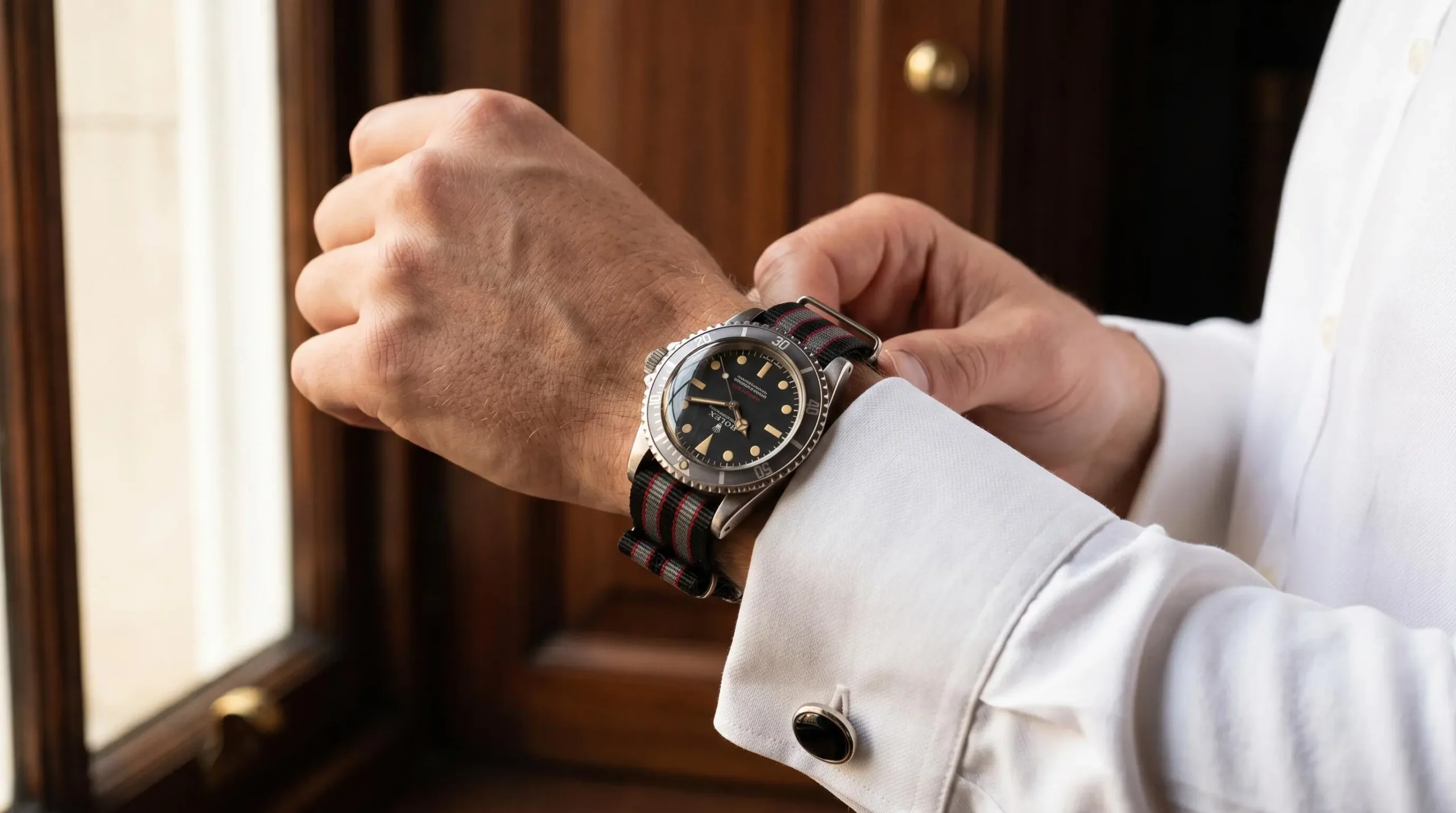 Gros plan d'un poignet masculin portant une Rolex Submariner vintage sur bracelet NATO rayé gris-noir-rouge, manchette de chemise blanche visible, main ajustant un bouton de manchette, intérieur élégant aux boiseries sombres en arrière-plan flou