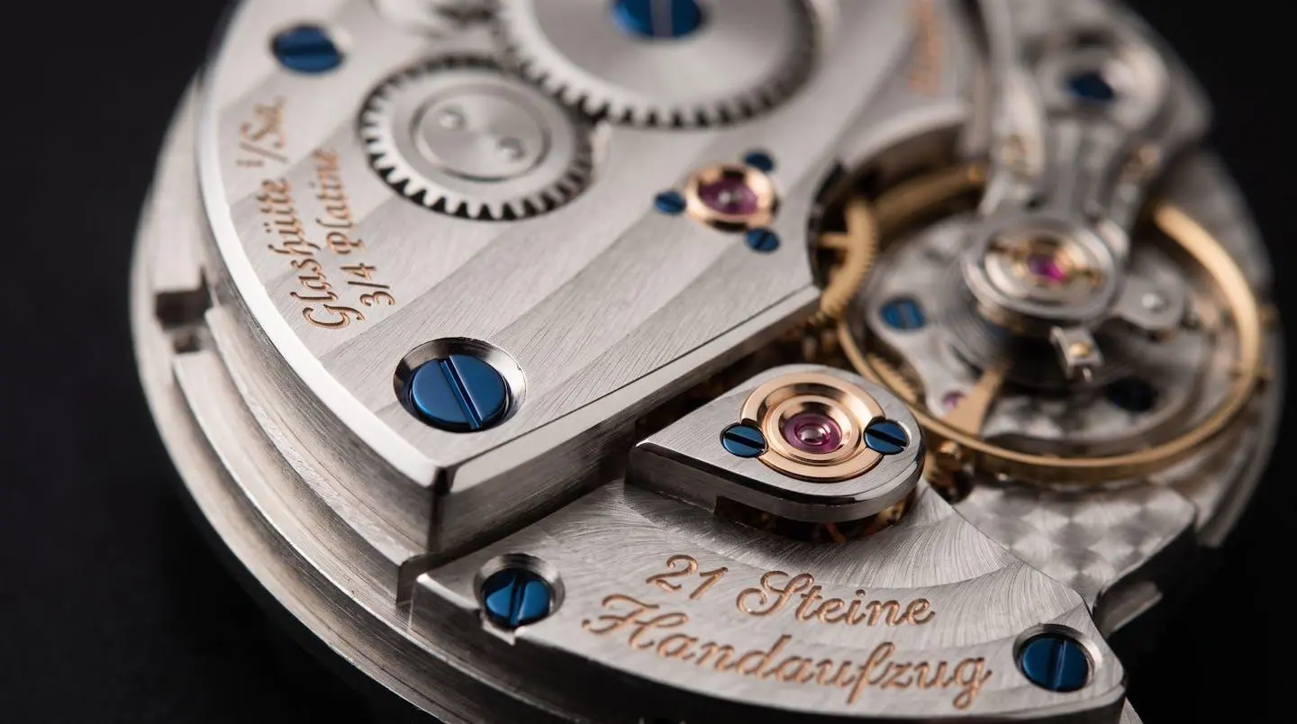Gros plan macro mouvement mécanique saxon, finitions haute horlogerie avec anglage miroir, perlage circulaire, vis bleues, gravure Glashütte, or rosé et acier poli, lumière rasante